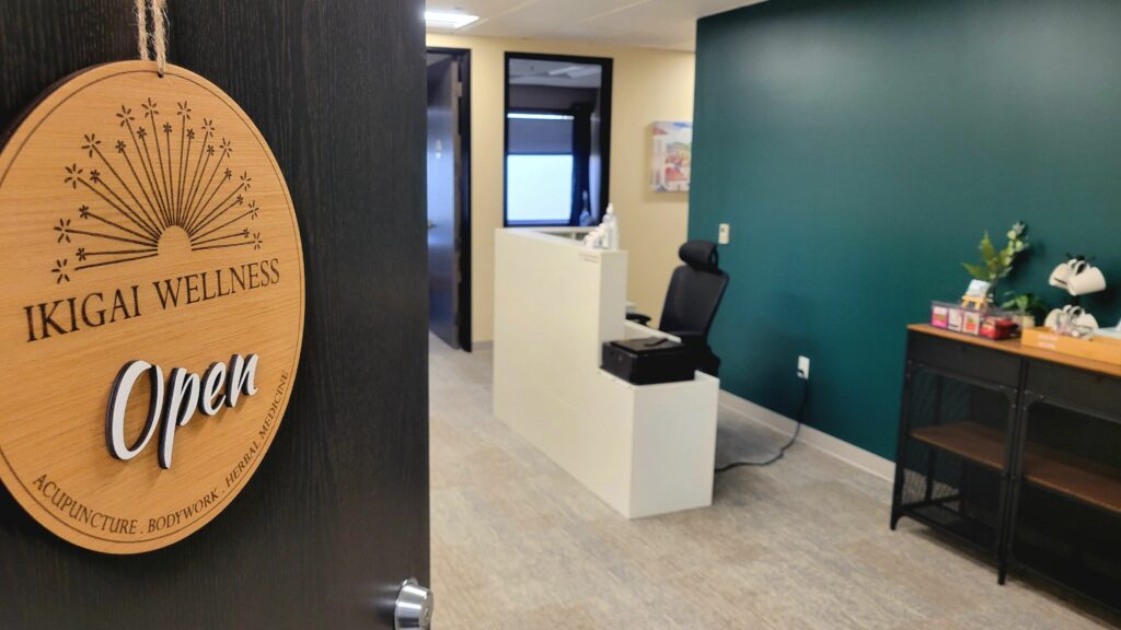 Photo of reception desk and tea station beyond an opening door with a wooden sign containing the Ikigai Wellness logo saying open.