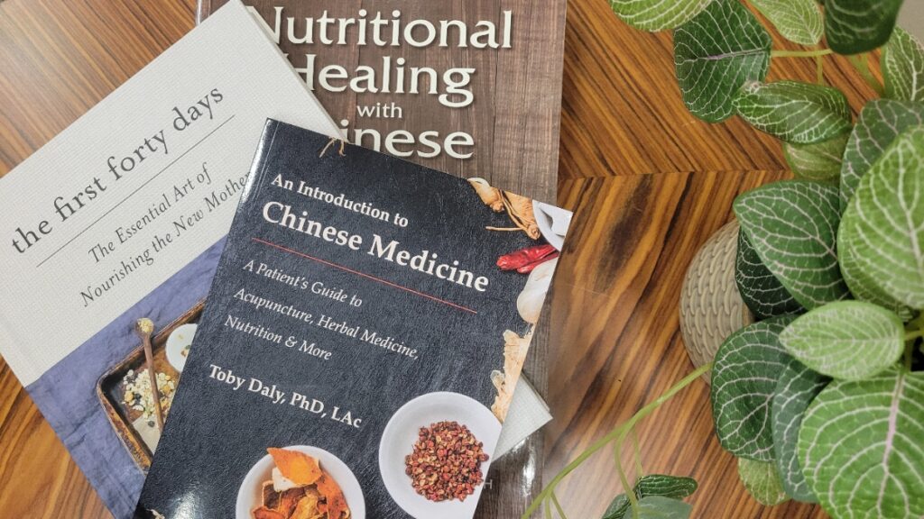Photo including the books titled "the first forty days", "an introduction to Chinese Medicine" and "nutritional healing with Chinese Medicine on a wooden background with a plant in the foreground.