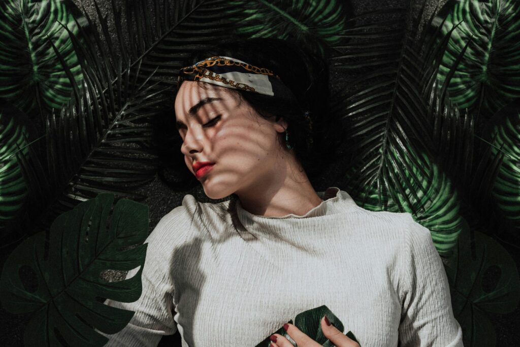 Woman laying down on a palm leaf background, her head turned to the left, eyes closed, hands to her chest.