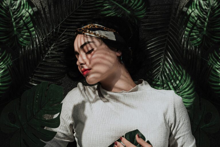 Woman laying down on a palm leaf background, her head turned to the left, eyes closed, hands to her chest.